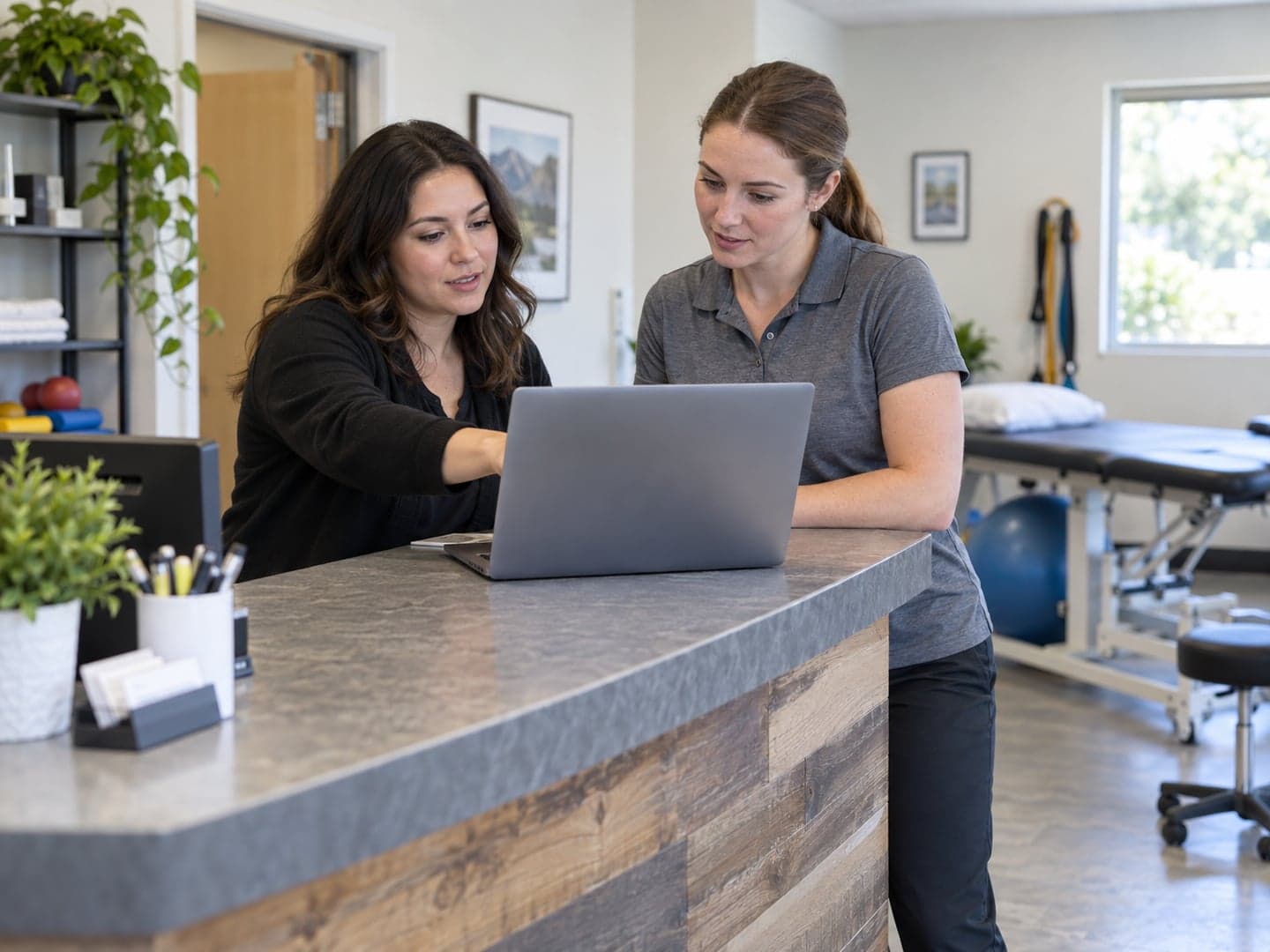 A front-desk coordinator and clinician reviewing a laptop together at a small physical therapy clinic
